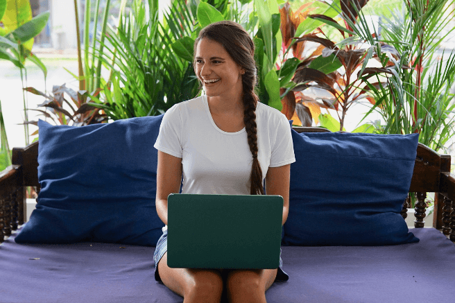 Nicola sits on a blue couch with her laptop on her lap. There is greenery behind her and she's smiling while looking to the side.